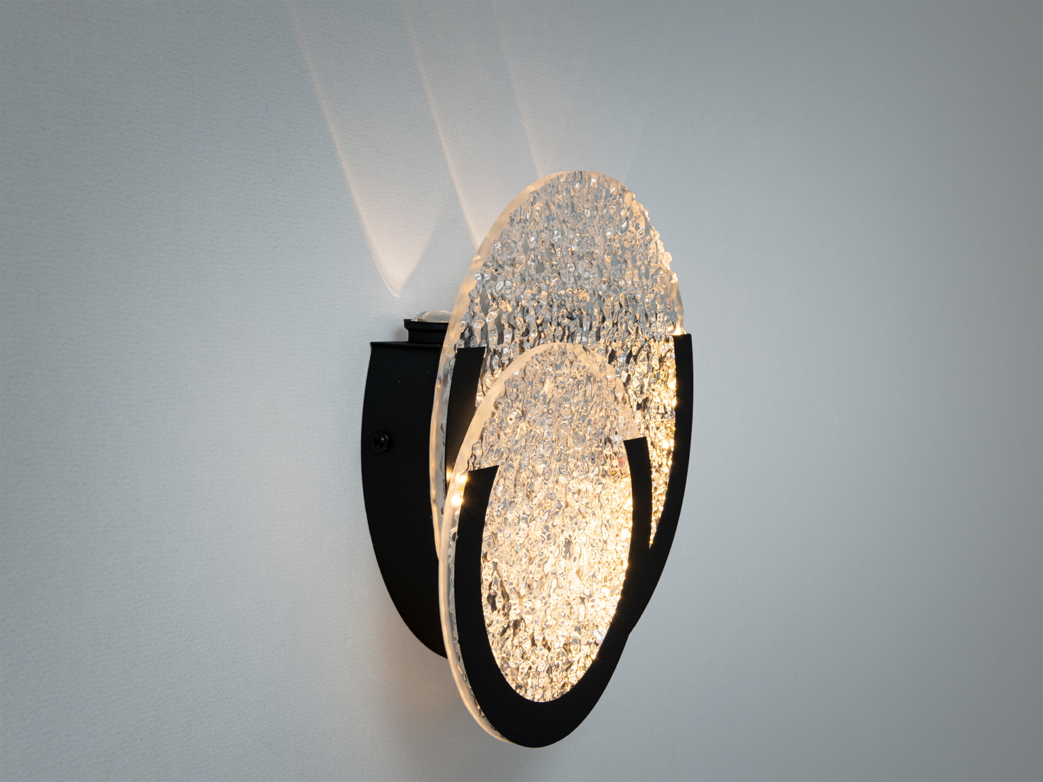 Дизайнерський світильник із прозорого текстурованого скла, що монтується на стіну. Чорні металеві елементи створюють контраст з білим фоном.